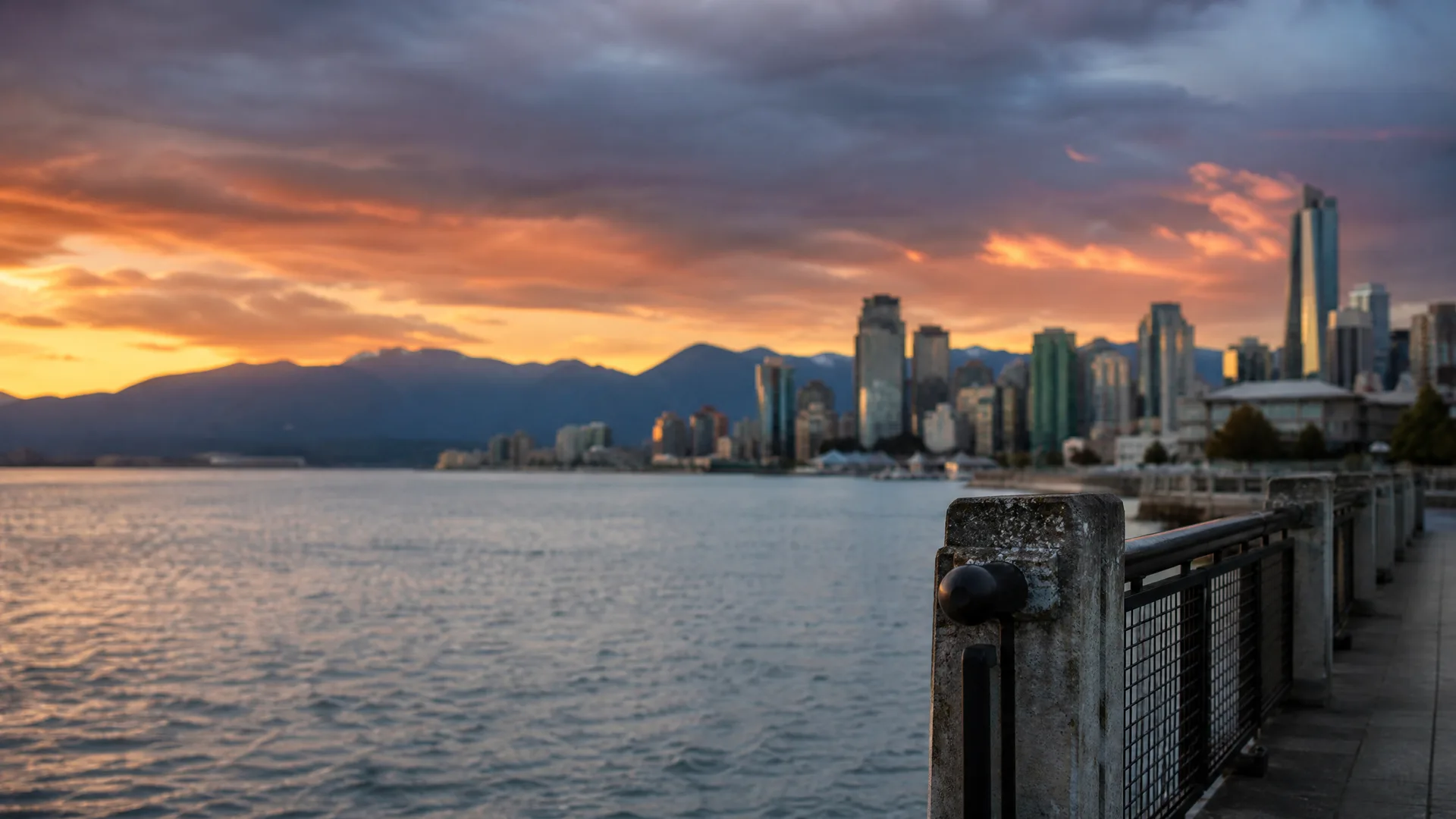 A waterfront seawall with a concrete post and metal railing in the foreground, looking across calm water toward a city skyline of high-rise towers and distant mountains under an orange and grey sunset sky.